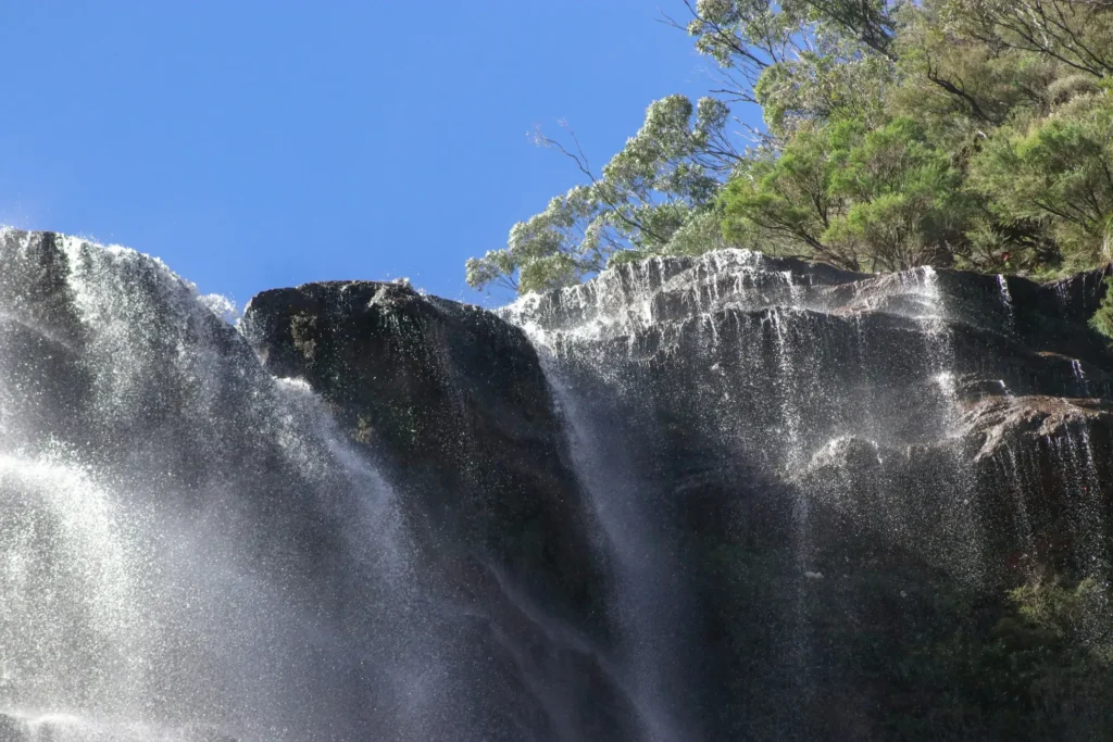 A low-angle view shows water cascading over dark, jagged rock tiers surrounded by lush green foliage. Sunlight catches the shimmering mist against a clear, vibrant blue sky.