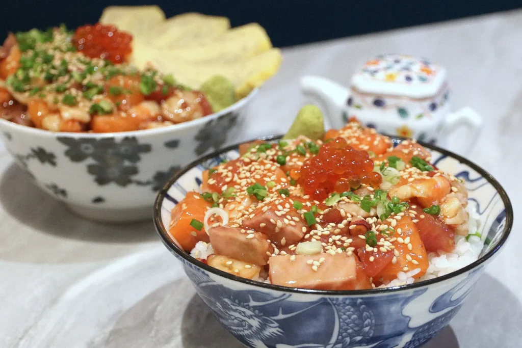 A detailed close-up of a Japanese chirashi bowl filled with white rice, cubes of fresh sashimi, salmon roe, chopped scallions, and sesame seeds. A second rice bowl and a small patterned ceramic teapot sit softly blurred in the background.