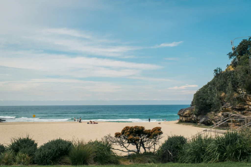 A sunny coastal beach with golden sand, soft ocean waves, and a rocky cliff bordering the right side. People gather along the shore under a bright blue sky filled with wispy white clouds.