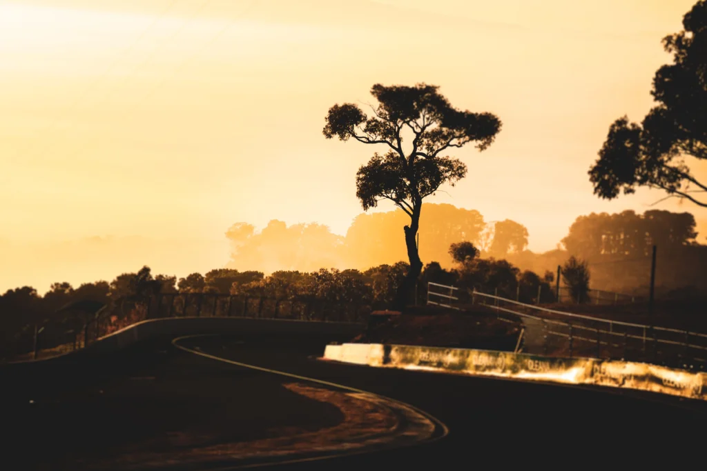 A winding asphalt racetrack curves through a rural landscape bathed in warm golden sunlight. Silhouetted trees and gentle hills frame the track under a hazy, brightly illuminated sky.