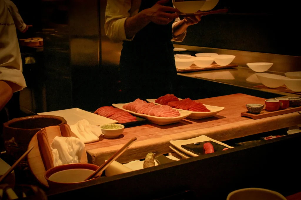 Plates of fresh, sliced raw tuna sit arranged on a wooden counter inside a dimly lit sushi restaurant. In the background, chefs in white coats and aprons prepare dishes and hold serving trays.