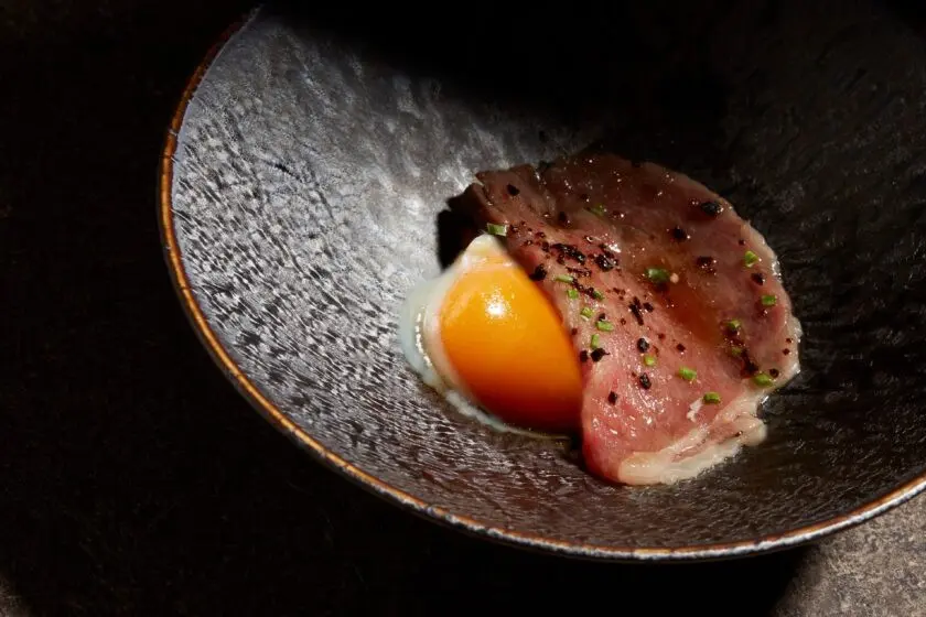 A thinly sliced piece of seared Miyazaki A5 Wagyu beef sits in a dark ceramic bowl alongside a soft-boiled egg yolk. The beef is garnished with finely chopped herbs and pepper, highlighted by ambient lighting against a dramatic black background.