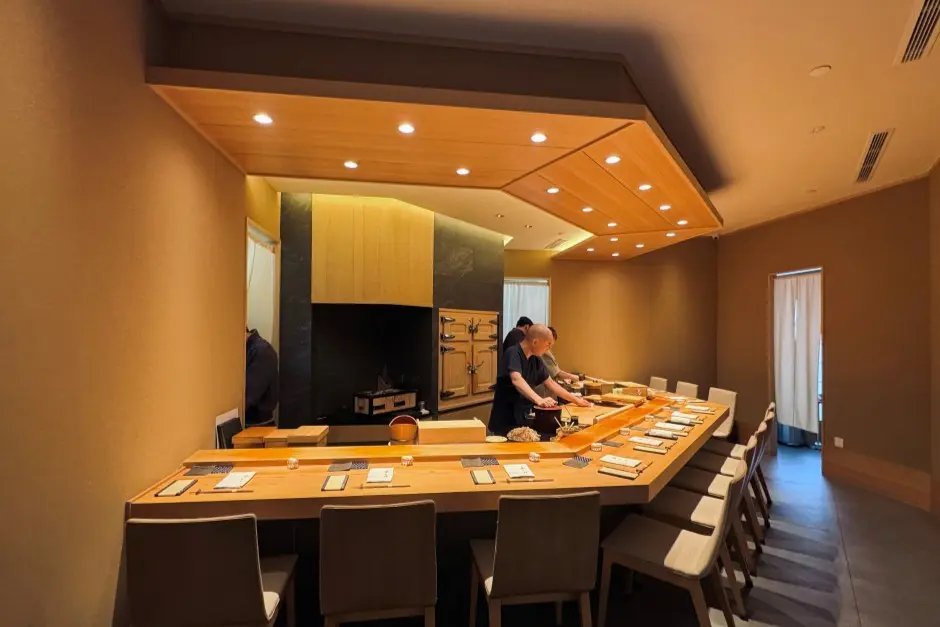A chef with a shaved head prepares the dining area behind an L-shaped wooden counter set with individual place settings in a modern Japanese restaurant. Warm overhead recessed lights illuminate the light wood paneling, minimalist beige walls, and neatly arranged seating.
