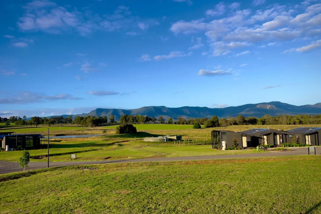 Modern, dark-colored cabins sit nestled within vast green lawns and rolling fields under a bright blue sky filled with light clouds. In the background, a distant mountain range stretches across the horizon beneath the open sky.