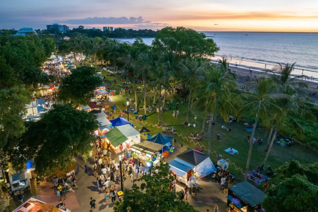 An aerial view shows a bustling outdoor market filled with food stalls, crowds of people, and tall palm trees along a grassy park adjacent to a sandy beach. In the background, a calm ocean stretches toward the horizon beneath a vibrant sunset sky filled with warm orange and yellow hues.