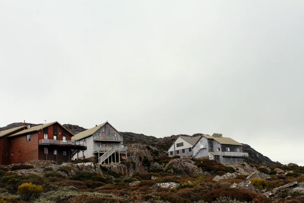 Several wooden and weathered grey lodges sit nestled along a rocky, brush-covered hillside. Overhead, a pale, overcast sky creates a muted, serene landscape.