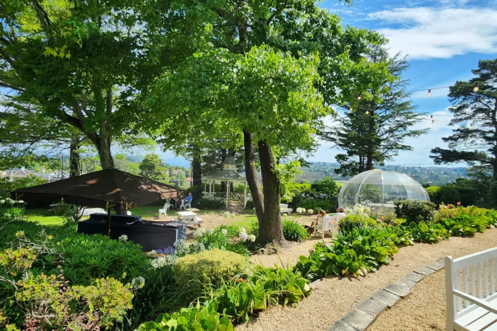 A sunlit, lush garden dining area features mature leafy trees, a white gazebo, and a clear dome dining Igloo. Outdoor seating, patio umbrellas, and lush greenery frame a scenic view of rolling hills in the background.