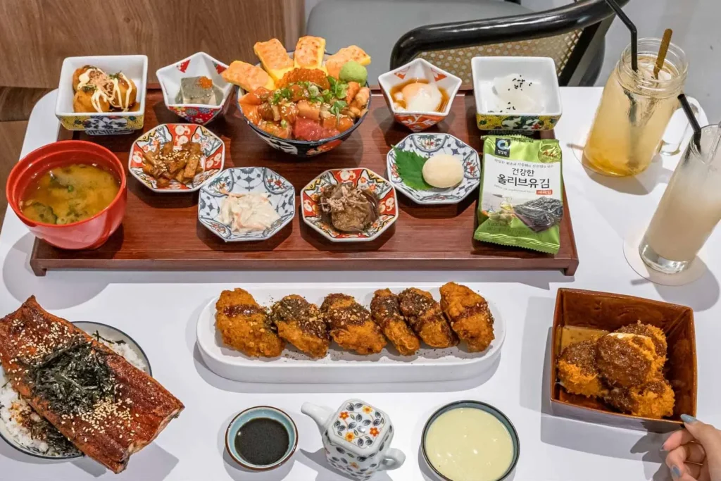 A lavish Japanese meal spread features a large set tray filled with a chirashi don bowl, tamagoyaki, miso soup, seaweed snack, and various small side dishes in decorative ceramic bowls. Surrounding the wooden tray on the table are additional dishes including grilled unagi over rice, fried chicken wings, katsu sauce, small condiment bowls, and iced drinks.