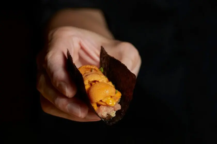 A hand holds a crisp nori seaweed wrap filled with minced tuna and generously topped with fresh orange sea urchin (uni). The gourmet sushi bite is presented up close against a dark, dramatic background.