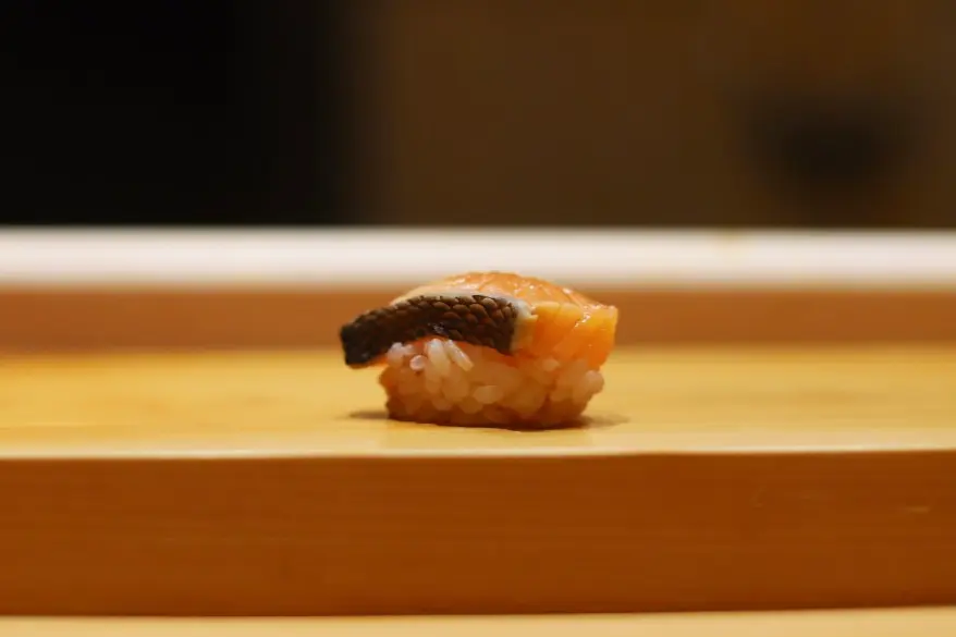 A single piece of salmon nigiri sushi with dark fish skin sits atop seasoned rice on a light wooden counter. The background is softly blurred in warm tones, emphasizing the clean presentation of the dish.