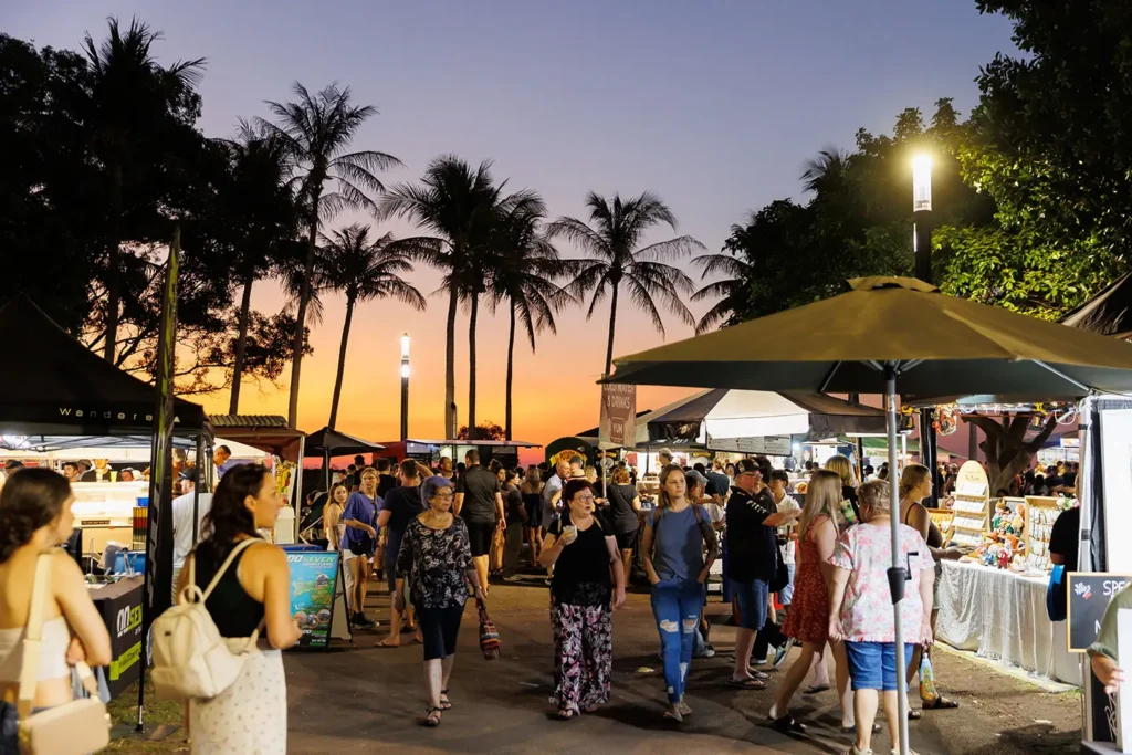 People stroll through a lively outdoor night market filled with food stalls, vendor tents, and illuminated streetlights. Silhouetted palm trees stand against a vibrant orange and purple sunset sky in the background.