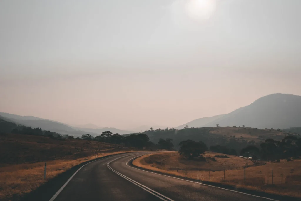A paved two-lane road winds through a dry, golden landscape flanked by low hills under a hazy sky. The sun shines softly above the hazy mountain ridge in the distance, illuminating the quiet countryside.
