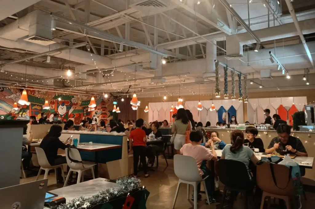 A crowded restaurant dining room features patrons seated at various tables under exposed ceiling beams and ductwork. Warm string lights and cone-shaped pendant lamps illuminate the space, which is decorated with festive garland and a colorful graphic mural on the back wall.
