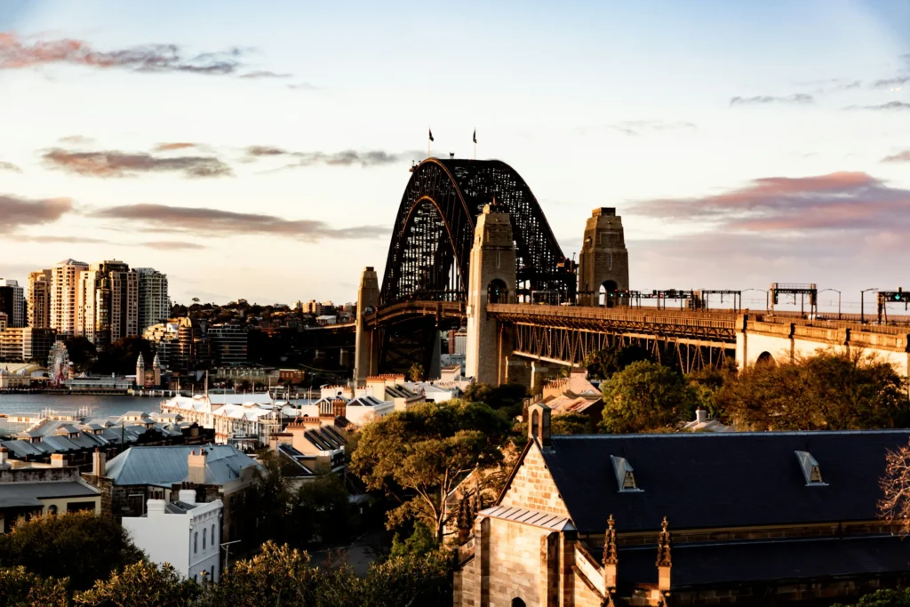 A scenic view of the Sydney Harbour Bridge towering over surrounding buildings and lush trees during a warm, bright twilight. Soft sunlight illuminates the cityscape, casting a glowing light across the water and sky.