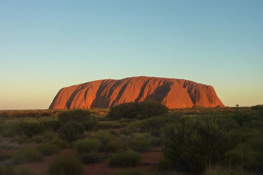 A massive, reddish sandstone rock formation glows under a warm golden light against a clear blue sky. Low green shrubs and dry desert vegetation scatter across the flat foreground landscape.