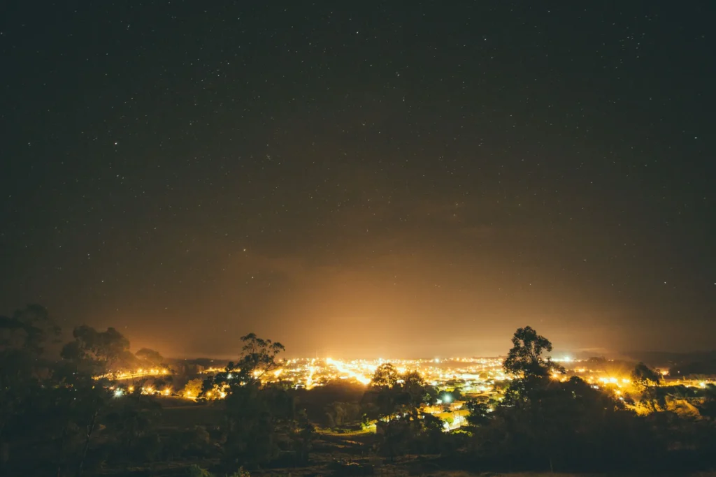 A panoramic long-exposure photograph captures a brightly lit town nestled beneath a dark sky filled with stars. Silhouetted trees frame the foreground, while a warm golden glow radiates from the city lights into the night atmosphere.