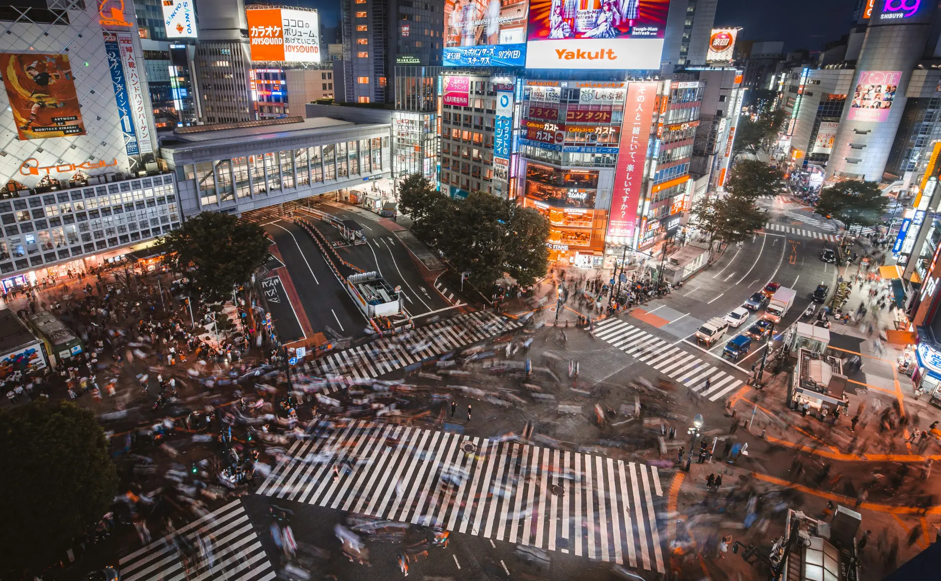 An elevated, long-exposure view of Shibuya Crossing in Tokyo glowing under bright neon advertisements at night. Blurred crowds of pedestrians stream across the iconic multi-directional crosswalks while surrounding city traffic moves through the intersection.