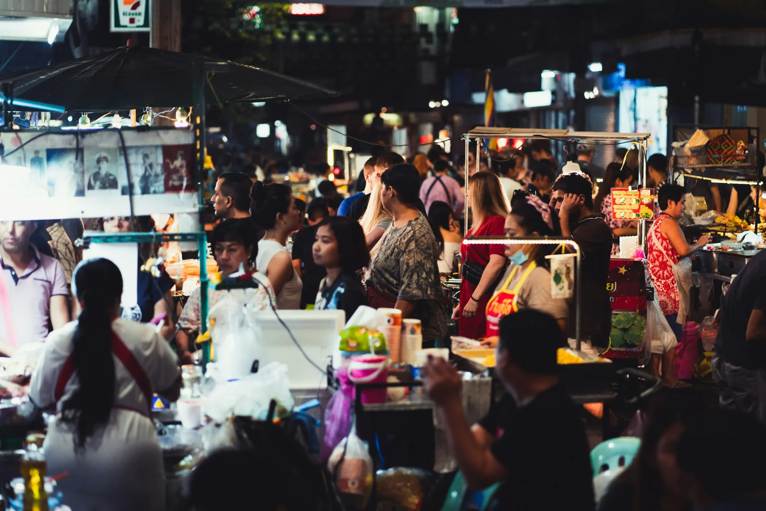 A crowded night market bustling with people browsing illuminated street food stalls. Vendors prepare dishes and serve customers under brightly lit outdoor carts.