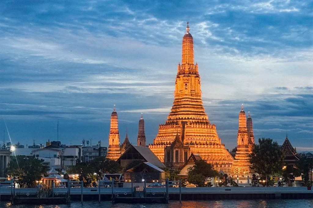 Wat Arun illuminated at dusk along the Chao Phraya River in Bangkok, with its central temple spire glowing against a blue evening sky.