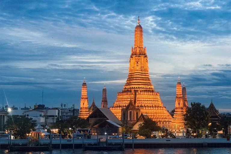 Wat Arun illuminated at dusk along the Chao Phraya River in Bangkok, with its central temple spire glowing against a blue evening sky.