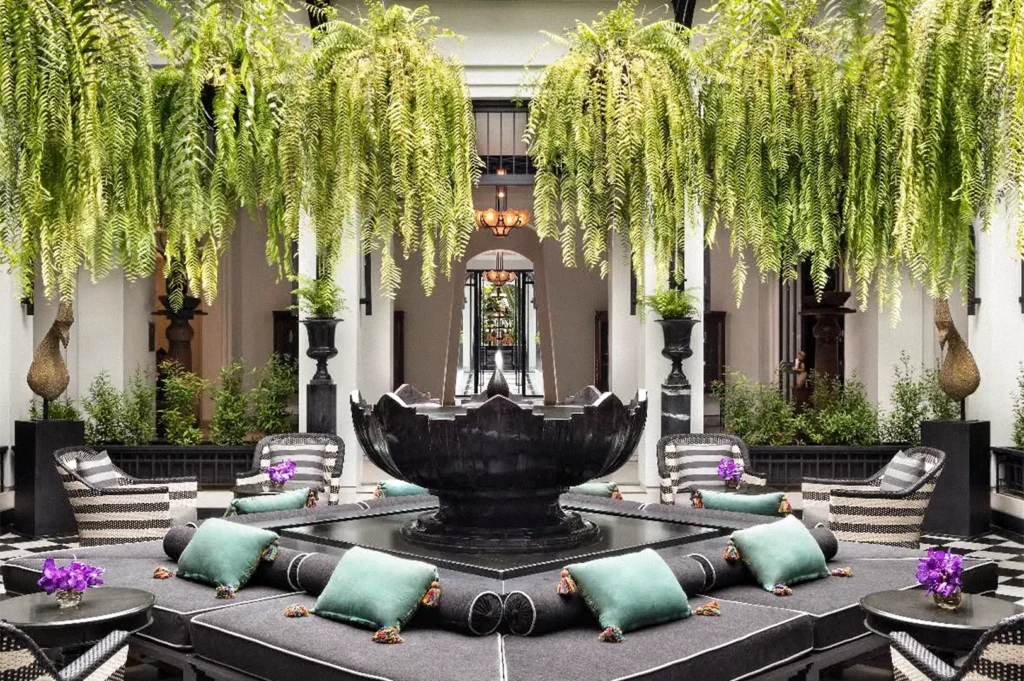 An elegant hotel courtyard with a large black fountain surrounded by cushioned seating, potted plants, and cascading ferns beneath white columns.