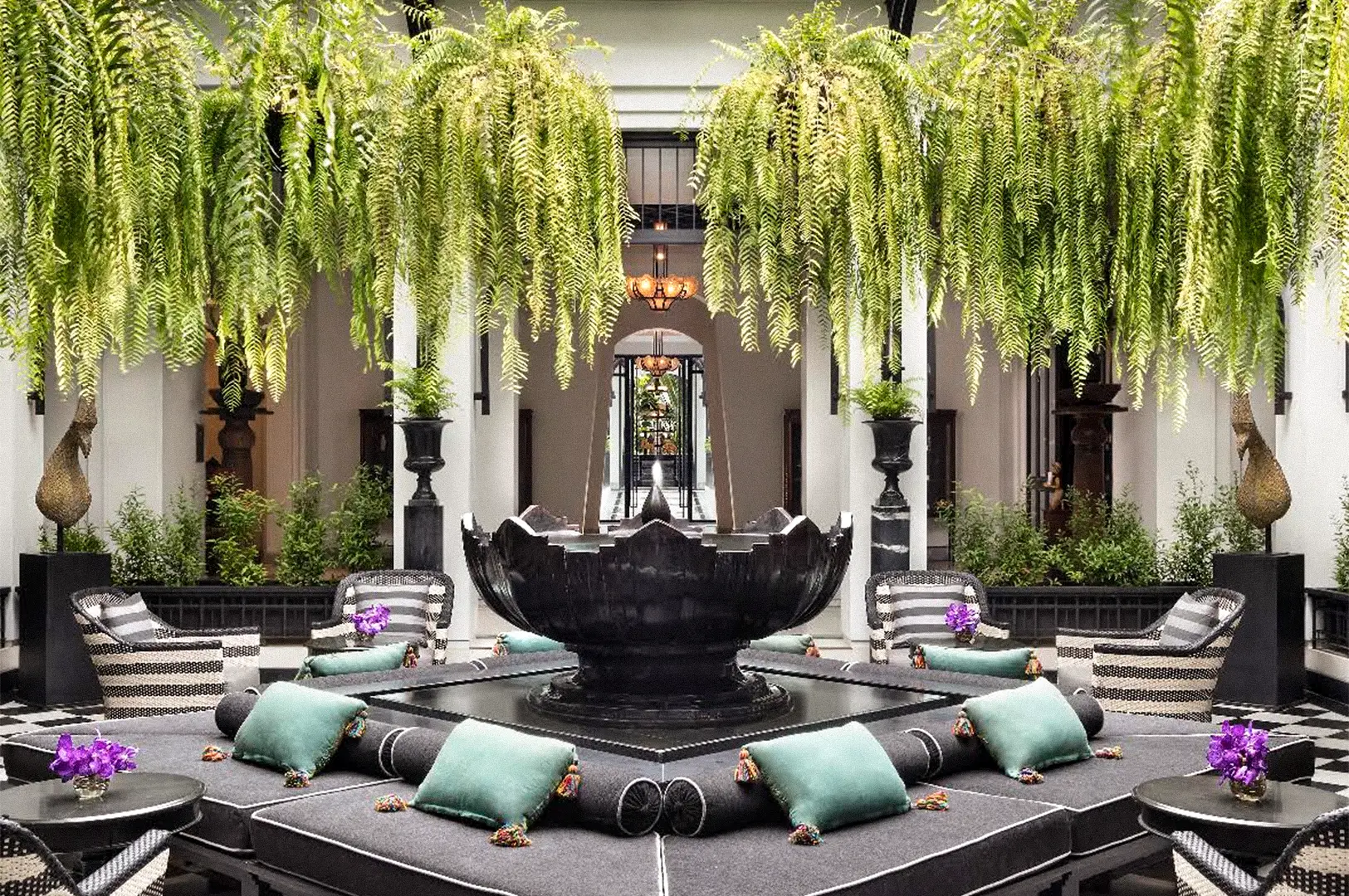 An elegant hotel courtyard with a large black fountain surrounded by cushioned seating, potted plants, and cascading ferns beneath white columns.