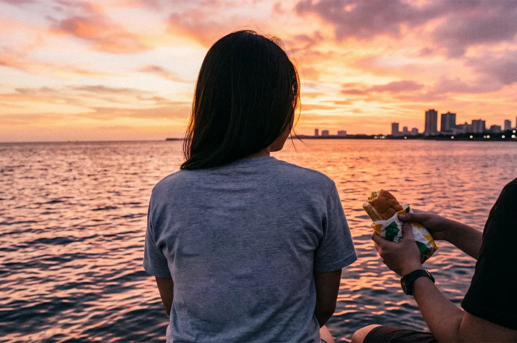 Two people sit by the water at sunset, with one person watching the orange-pink sky while the other holds a wrapped sandwich.