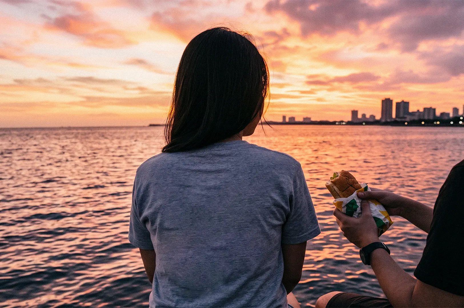Two people sit by the water at sunset, with one person watching the orange-pink sky while the other holds a wrapped sandwich.