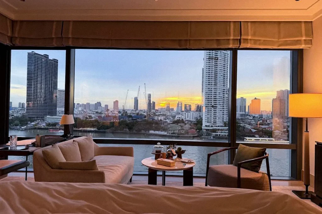 Luxury hotel room with floor-to-ceiling windows overlooking the Chao Phraya River and Bangkok skyline at sunset, furnished with a sofa, armchair, and coffee table.