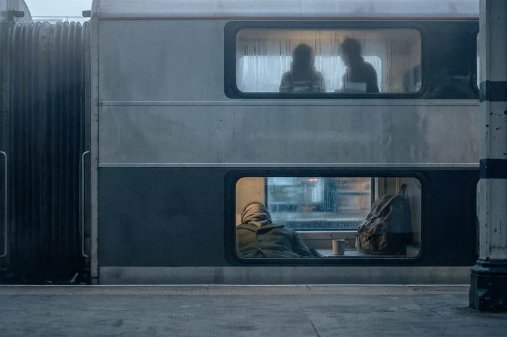 Exterior view of a double-decker overnight train carriage through a station window, showing a resting passenger and backpack in the lower compartment and two silhouetted passengers above.