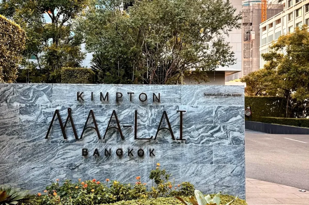 Exterior sign for Kimpton Maa-Lai Bangkok set against landscaped greenery and the hotel building.