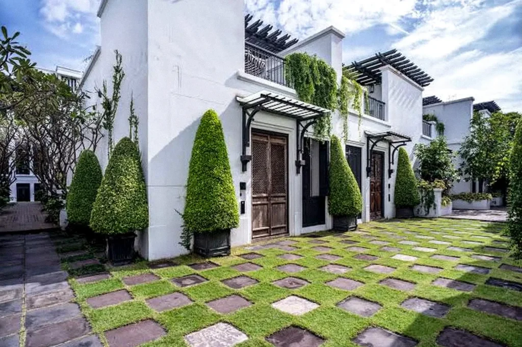 A bright white hotel courtyard with manicured cone-shaped trees, wooden doors, balconies, and a lawn of square stone pavers.