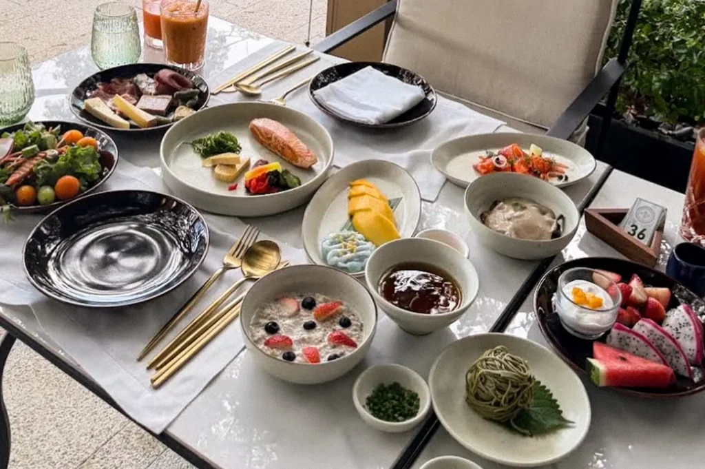Table filled with a variety of colorful dishes, desserts, fruits, and drinks at a dining setting overlooking the hotel grounds.
