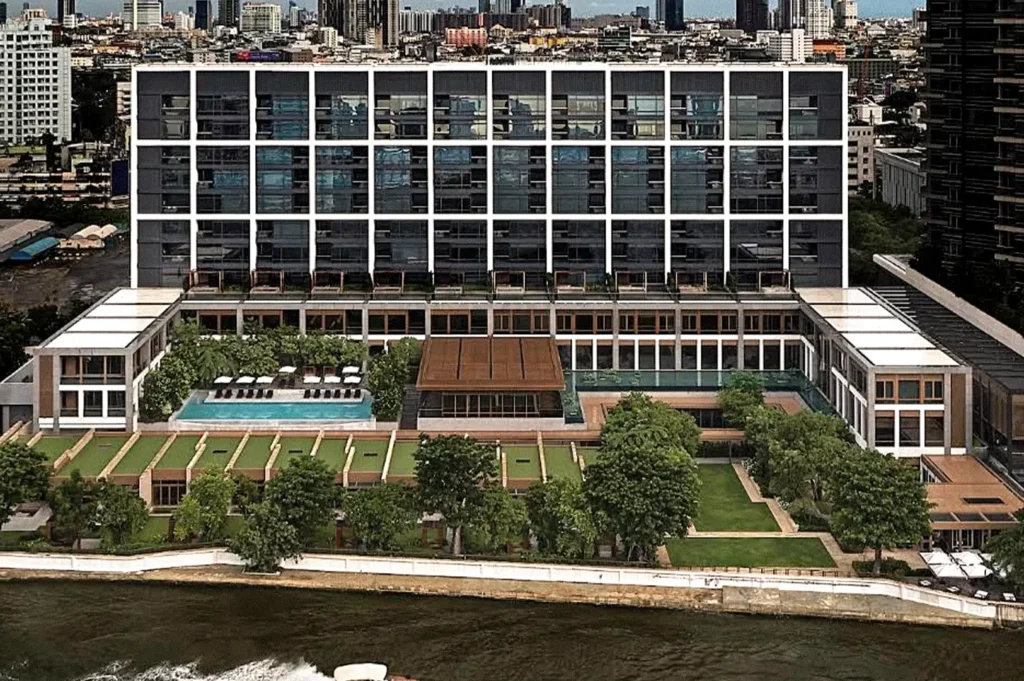 Aerial view of a modern riverside hotel in Bangkok, featuring a landscaped courtyard, swimming pool, and the Chao Phraya River in the foreground.