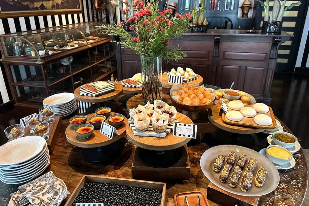 A hotel dessert buffet displaying assorted pastries, cakes, macarons, and plated sweets on wooden serving stands, with plates and utensils arranged nearby.
