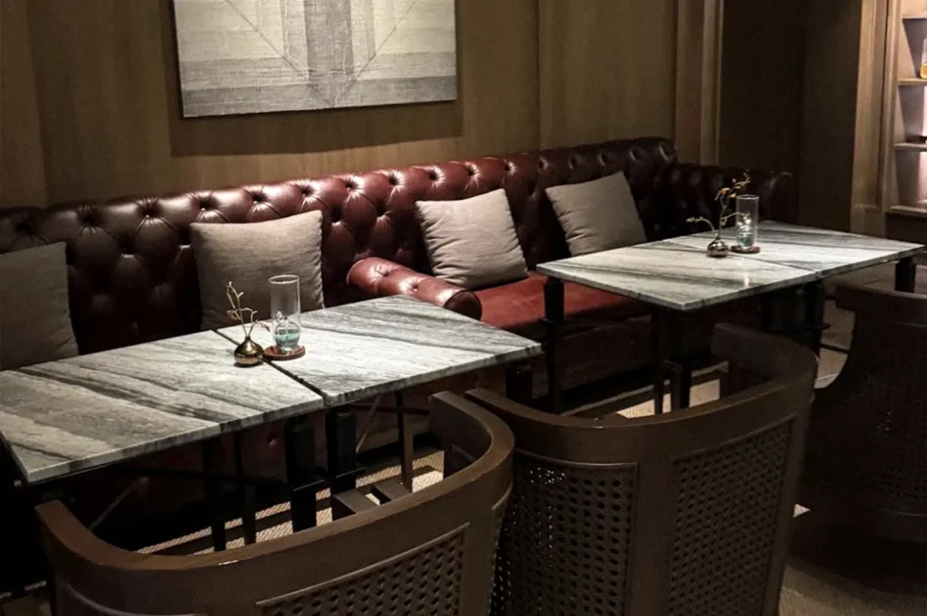 Dimly lit restaurant interior with marble-top tables, wooden chairs, and a long tufted leather banquette creating an intimate dining atmosphere.