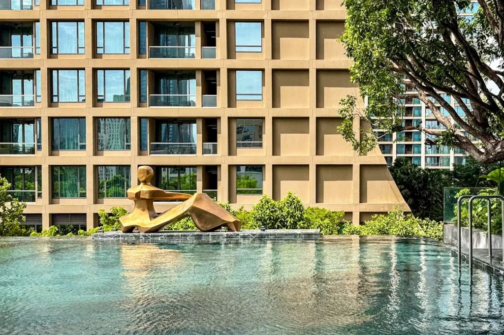 Outdoor hotel swimming pool with a bronze reclining sculpture, surrounded by greenery and a modern building.