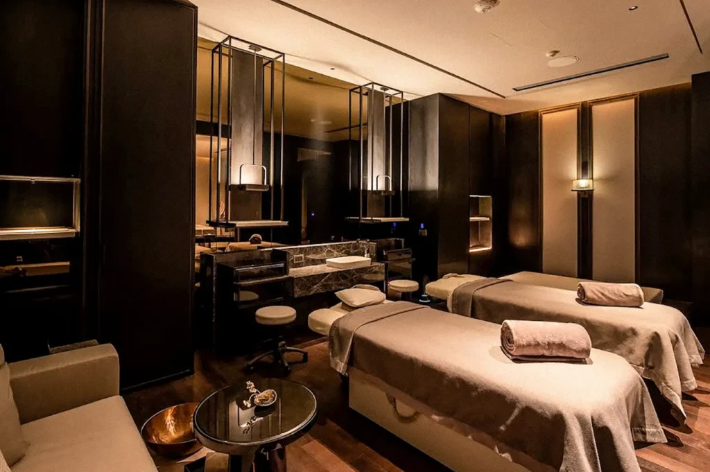 Luxury spa treatment room with two massage beds, warm ambient lighting, dark wood furnishings, and a marble treatment counter.