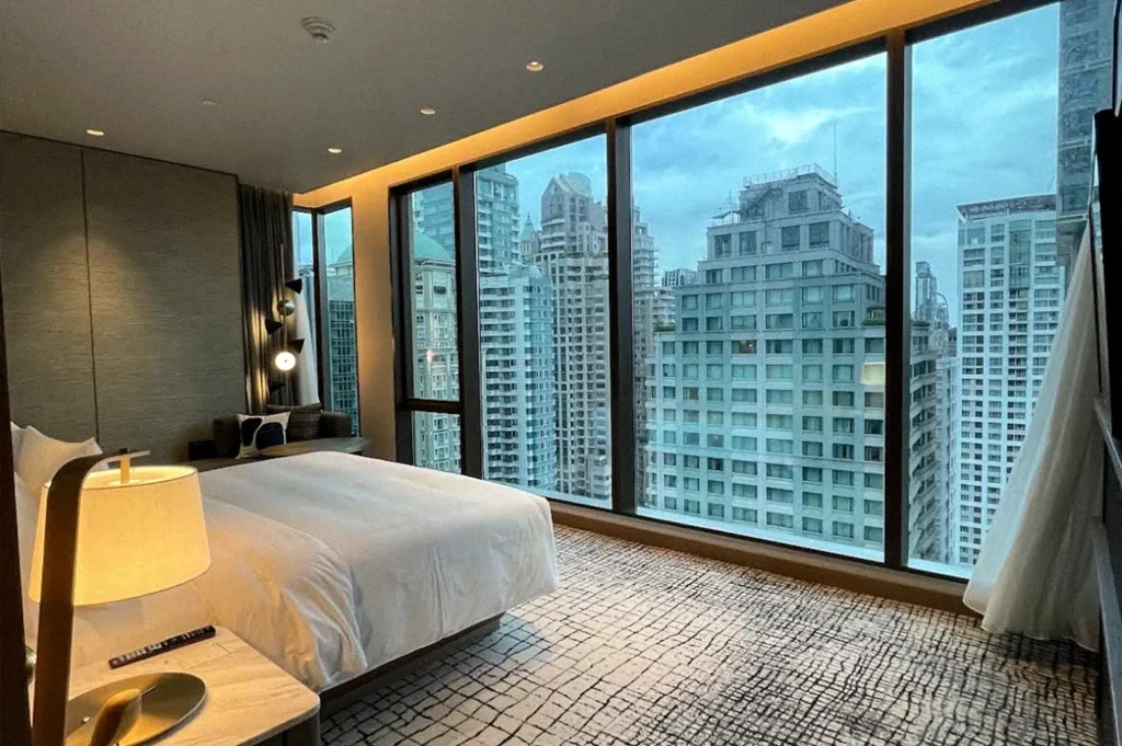 Kimpton Maa-Lai Bangkok guest room with a large bed, warm lighting, and floor-to-ceiling windows overlooking Bangkok’s high-rise skyline.