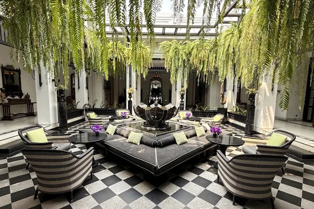 Elegant hotel courtyard with hanging ferns, black-and-white checkered flooring, woven lounge chairs, and a central decorative fountain.