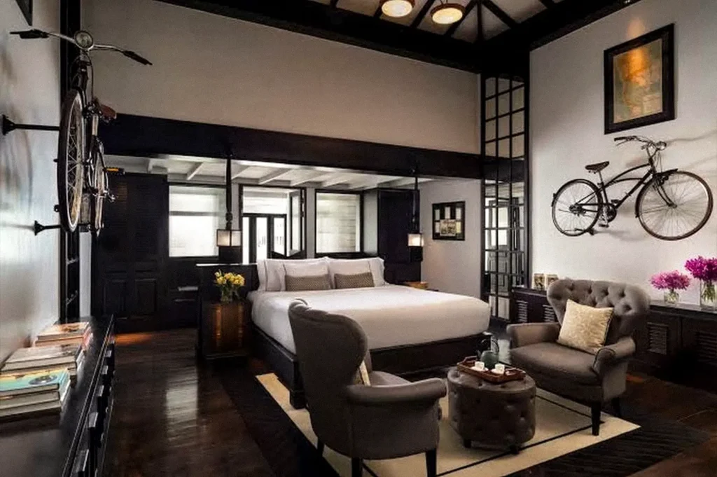 A sophisticated hotel bedroom with a large white bed, dark wood furnishings, two armchairs, a tufted sofa, and bicycles displayed as wall decor.
