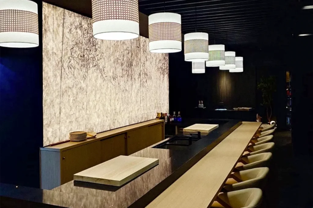 Elegant omakase restaurant interior with a long sushi counter, wooden seating, and patterned pendant lights.