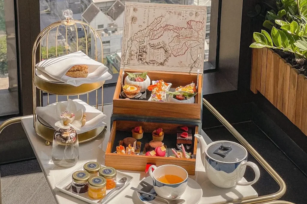 Hotel breakfast or afternoon tea spread arranged on a serving cart, featuring assorted pastries, savory bites, preserves, and tea beside a city view.