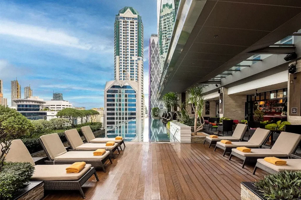 Rooftop hotel pool surrounded by lounge chairs and tropical greenery, with modern high-rise buildings visible across the Bangkok skyline.