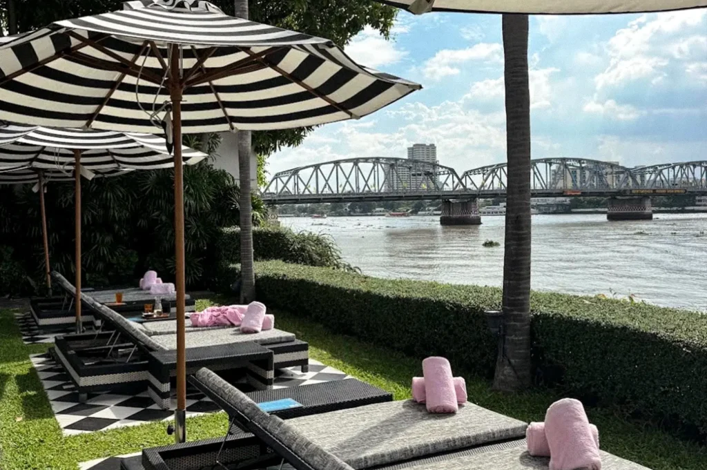 A riverside outdoor lounge with black-and-white striped umbrellas, cushioned sun loungers, palm trees, and views of a steel bridge across the river.