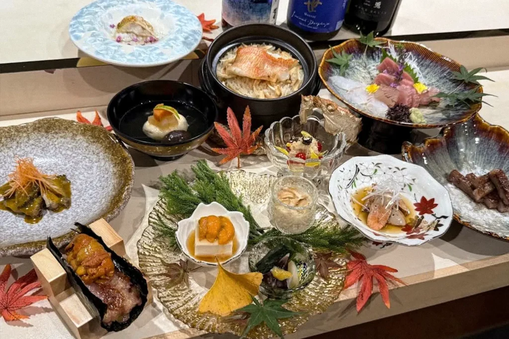 Assorted Japanese omakase dishes arranged on a table, including sashimi, grilled seafood, rice, and small seasonal appetizers garnished with maple leaves and flowers.