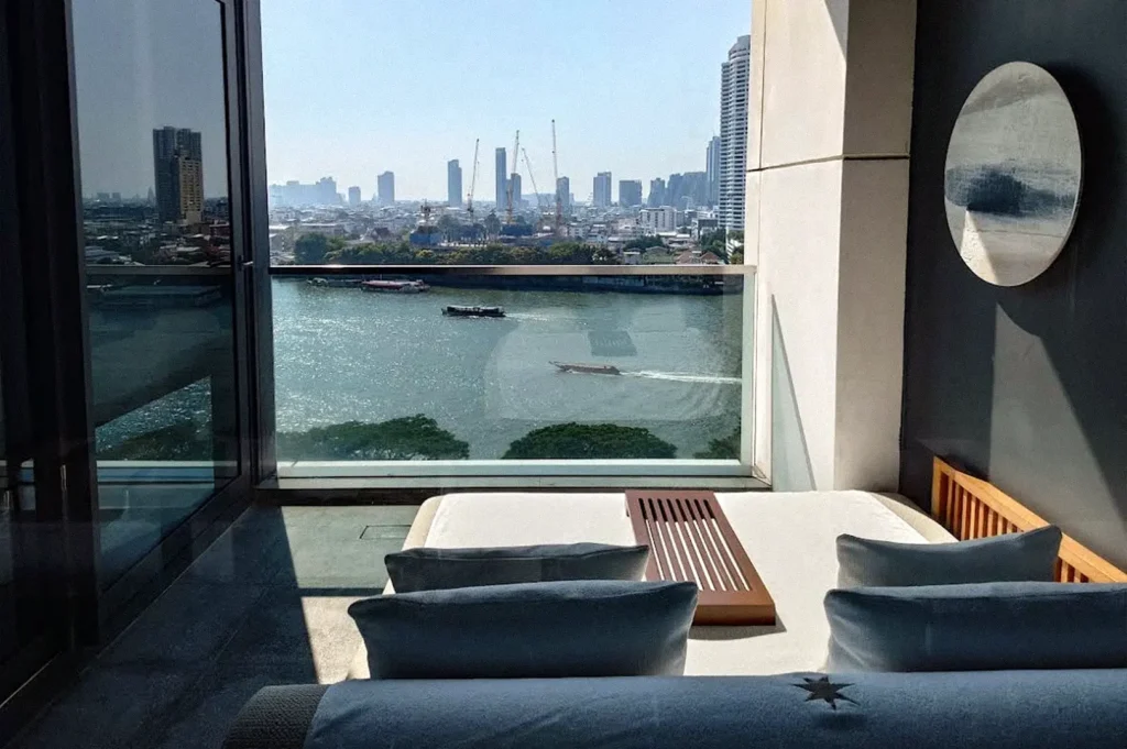 Hotel room with a large window overlooking the Chao Phraya River and Bangkok skyline, featuring a cushioned daybed and wooden table.