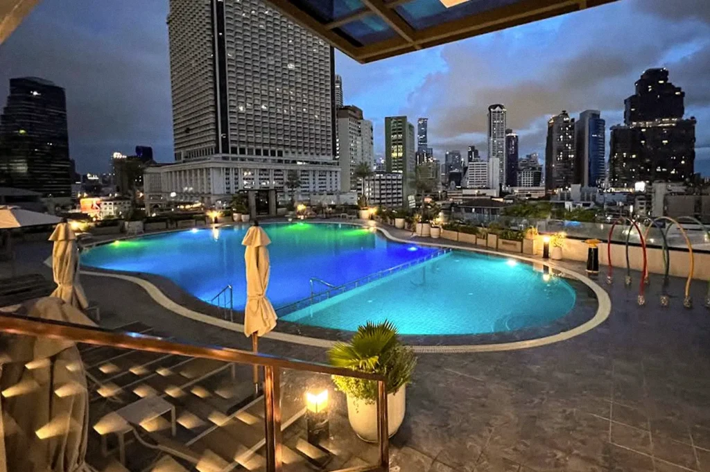 Rooftop hotel swimming pool glowing with blue and green lights at dusk, overlooking Bangkok’s illuminated city skyline.