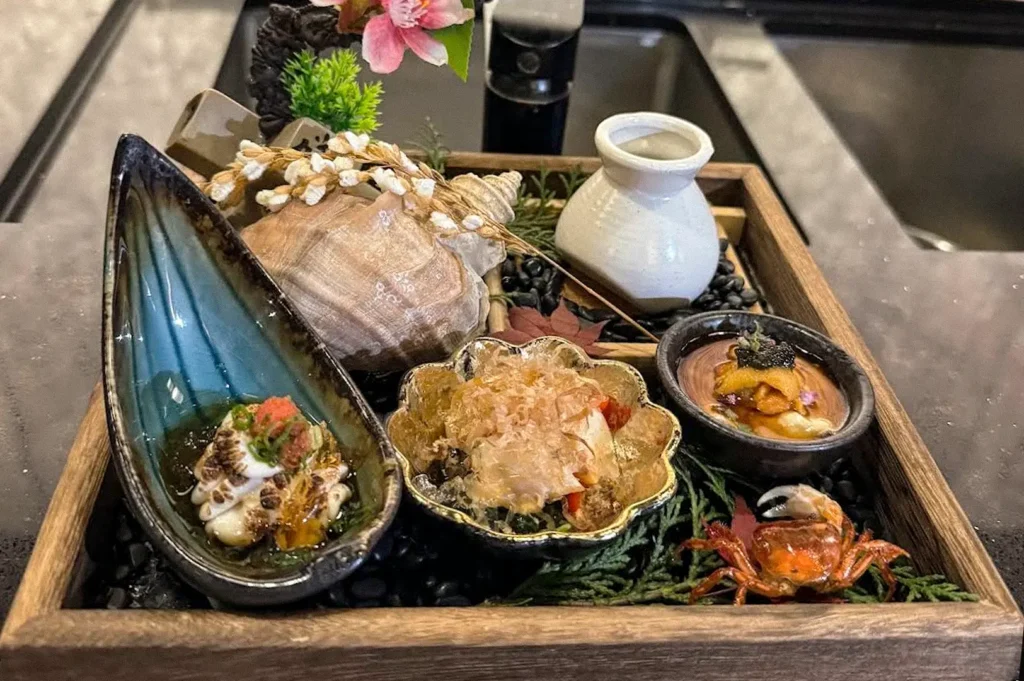 Japanese seafood appetizer platter featuring shellfish, sashimi, roe, and other delicacies arranged in decorative dishes on a wooden tray.