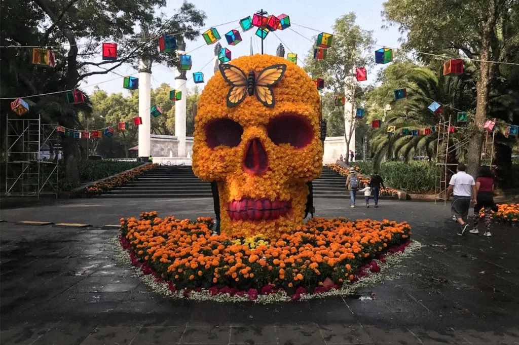 Large skull sculpture made from orange marigold flowers, decorated with a butterfly, surrounded by flower beds and colorful hanging lanterns in an outdoor garden.